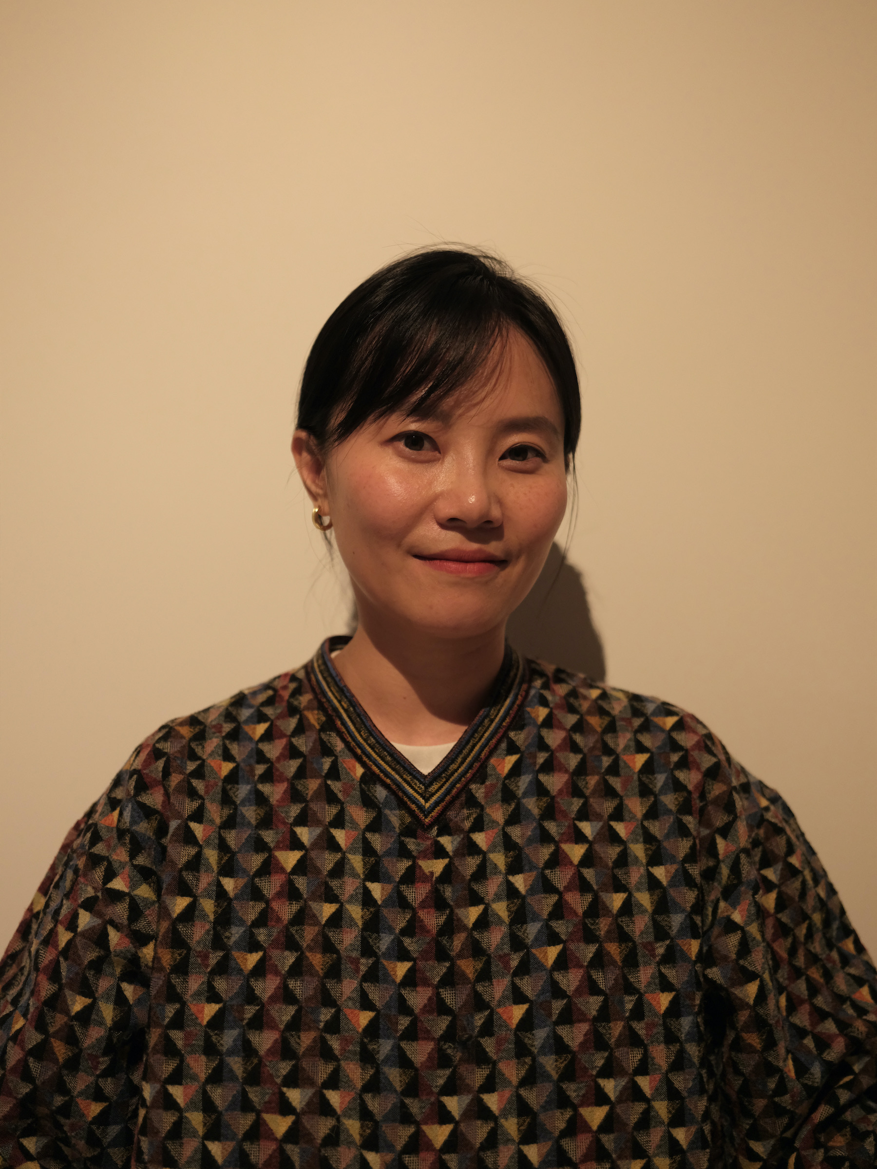 Yao, a Chinese woman with black hair, wearing a checkered sweater and smiling in front of a blank wall.